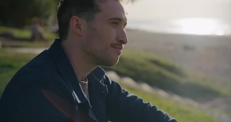 Man with a shirt and jeans sitting on hill slope uses smartphone next to the beach