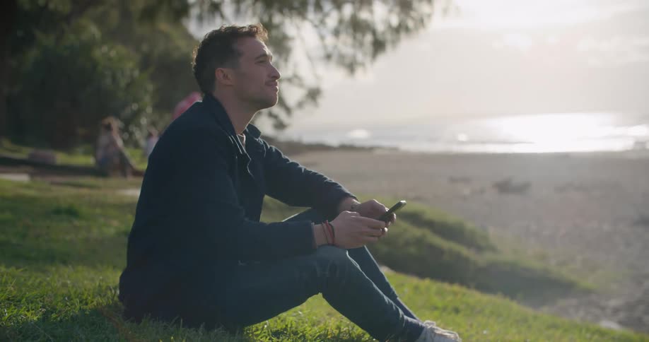 Man sits on grass near beach as waves roll in and scrolls through mobile phone