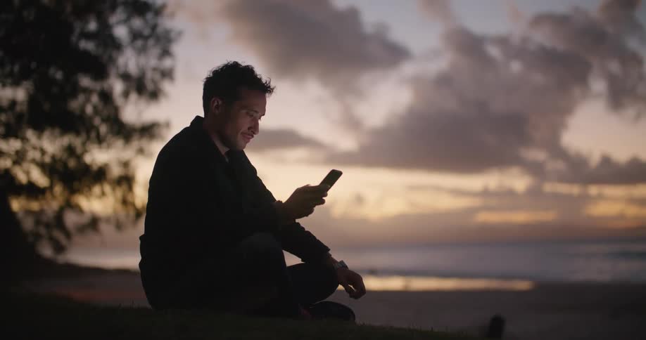 Man sitting cross legged on the ground uses smartphone on the beach at dusk