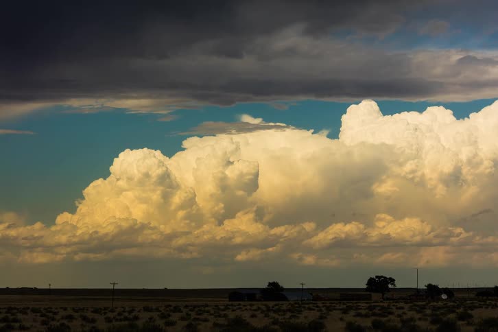 White clouds burst and expand in the blue sky as gray storm clouds cover the farm land