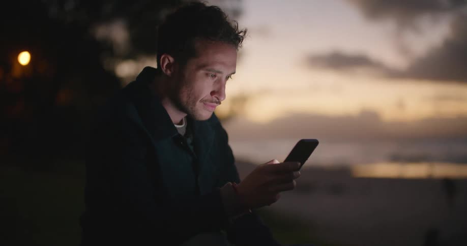 Man sits near beach as waves roll in viewing mobile phone with red sky on horizon
