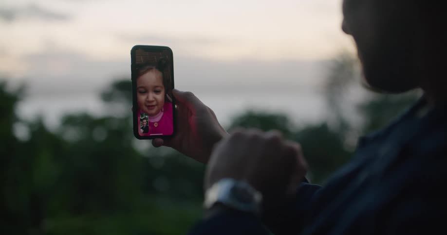 Father holding up the smartphone makes a video call with his daughter and is silhouetted at dusk outdoors