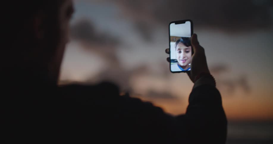 Man partly silhouetted against red sky talks via video call with boy on mobile phone