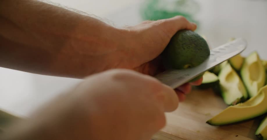Person slices avocado holding it in air next to avocado slices sitting on wooden chopping board