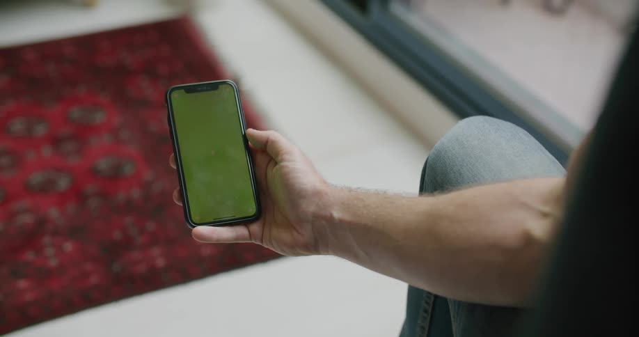 Man with jeans sits on couch and uses iphone smartphone against the red rug in the living room