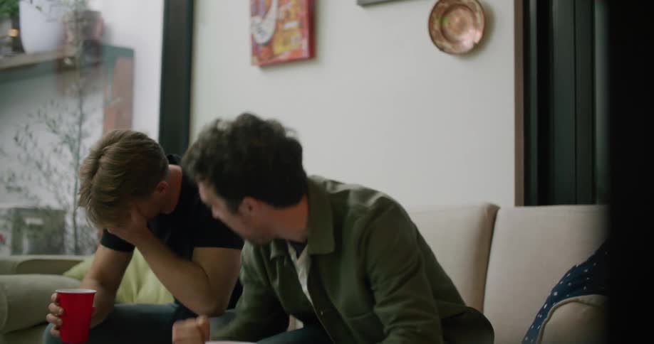 Two men in a room sitting on a couch with a wall and a window in the background