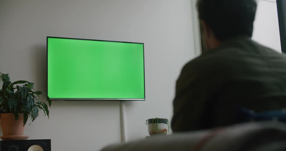 A man sits on the couch in front of a green screen television and pumps his arm in excitement