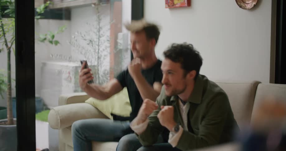 Two men in a room sitting on a couch with one holding a smart phone with a wall and window in the background