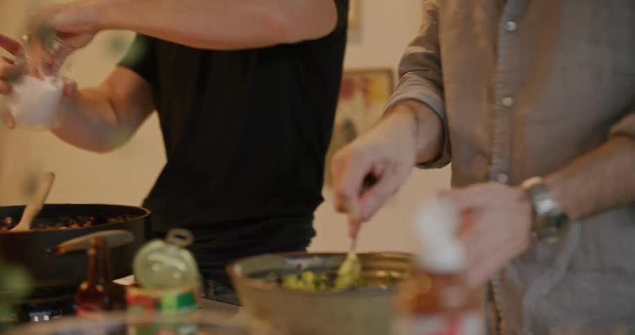 Two men work in the home kitchen beside each other tending to food in a pan and food in a bowl