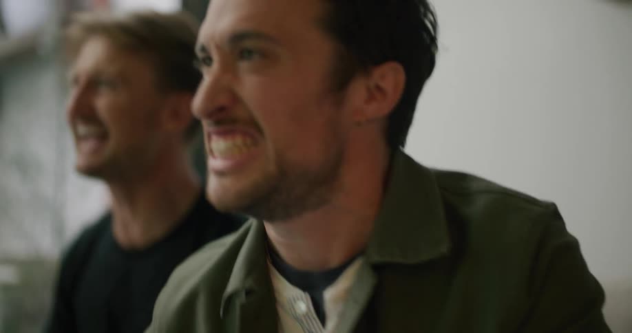 Two men sit on a couch and cheer and tap their glasses together