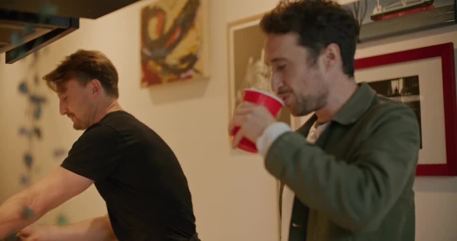 Men talk while one cooks over stove and the other takes a drink