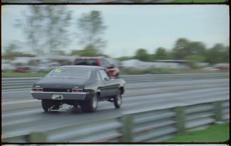 A red pickup truck and a black coupe race on a track as people watch