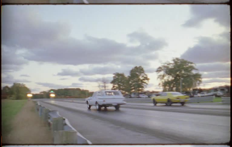 Cars move along road past parking lot and low buildings