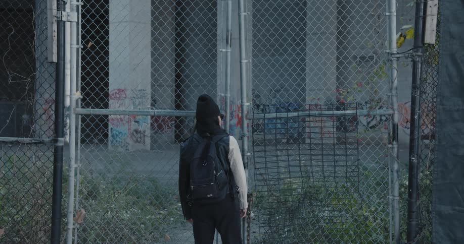 Woman stands near fence around graffiti covered building with overgrown yard