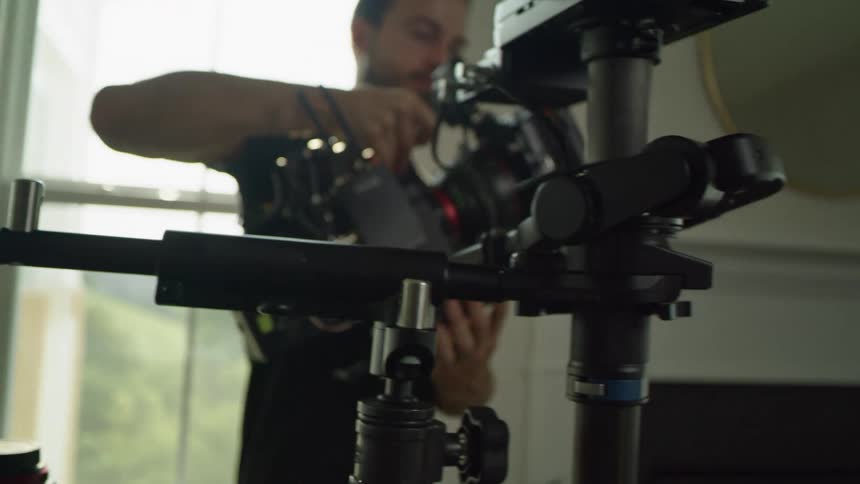 A man in a room putting a camera on a stand with a wall and window in the background