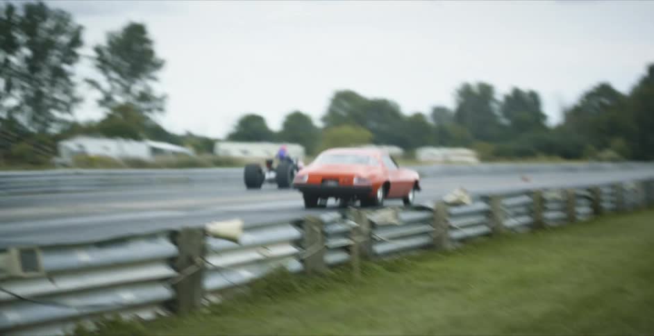 Two vehicles drag racing on a track with trees the sky and clouds in the background