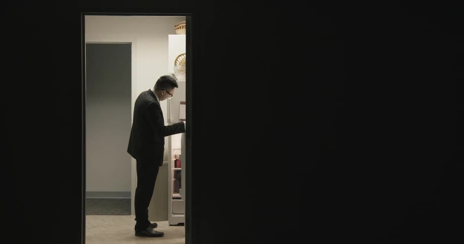 A man with glasses entering into a room and turning on the lights and opening the fridge