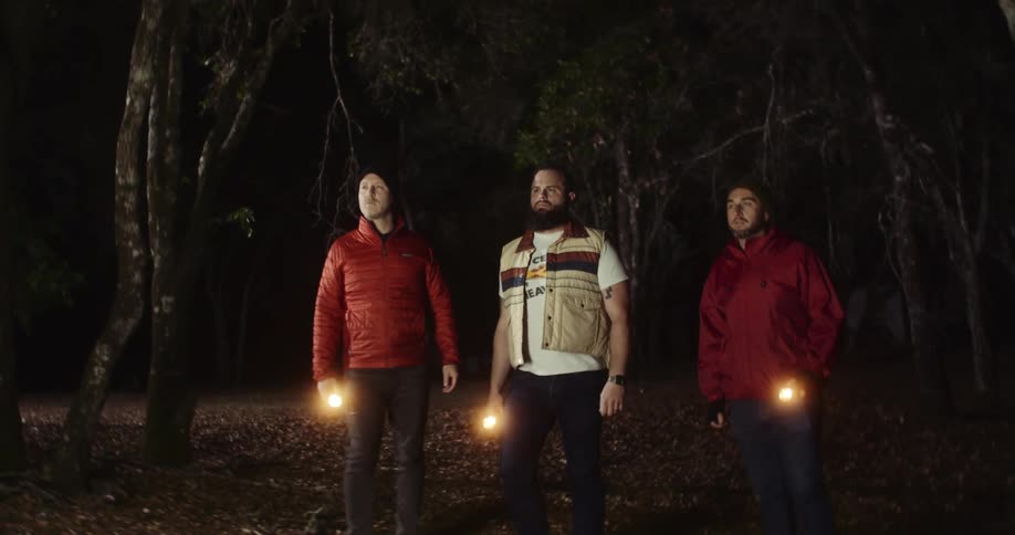Three men stand in a forest with flashlights and look at a lit up board