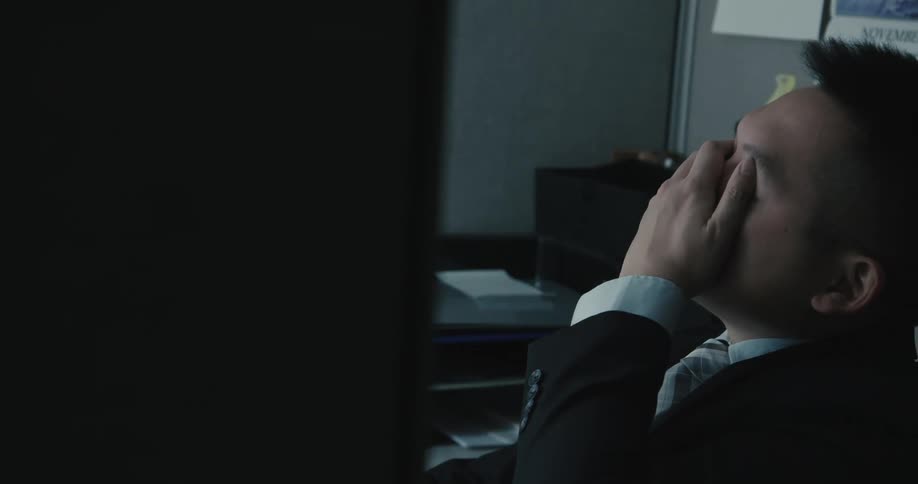 A man in an office leans back in his chair and takes his glasses off and rubs his eyes