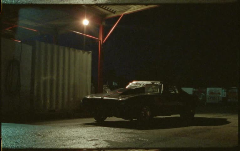Sports car sits parked near building under roof overhang with single light casting shadow on asphalt