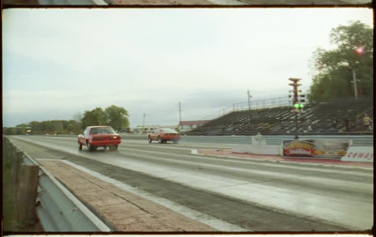 Two vehicles drag racing on a track with trees the sky and clouds in the background