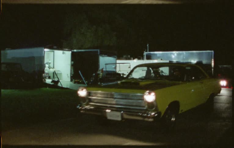 People move around among trailers as sports car drives down road nearby