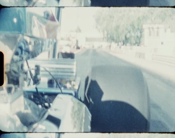 A go kart races down a straight road as a small crowd crowd watches from the sidewalk