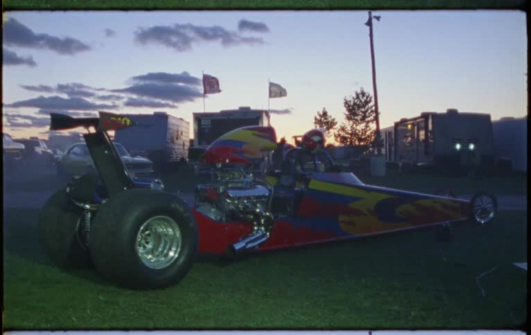 A drag racer sits on the track grass as the wind blows flags and trees