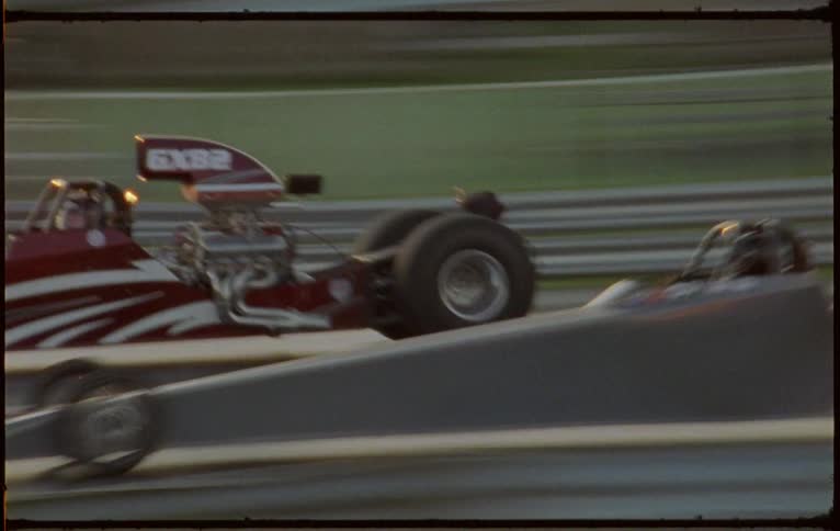 Two racers wearing race suits race race cars down track past foliage covered woods