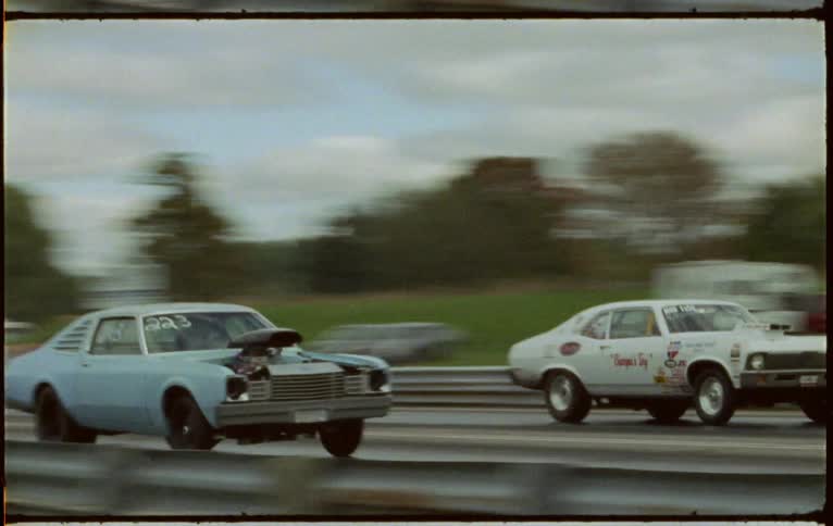 Two vehicles drag racing on a track with trees the sky and clouds in the background