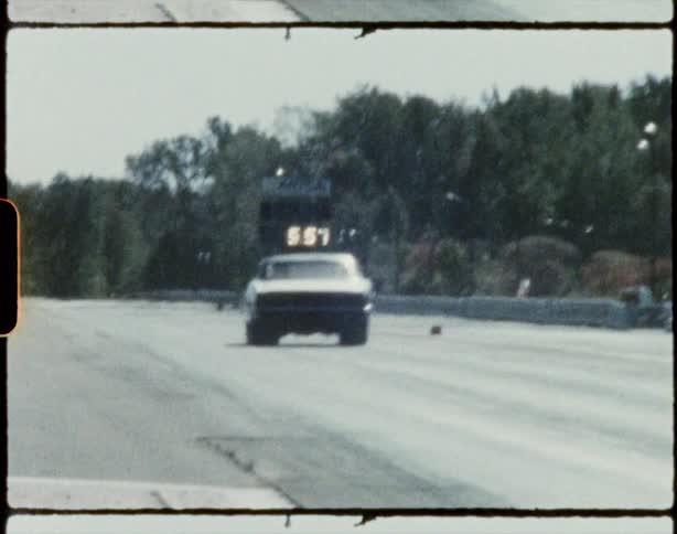 Race cars speed down track passing by woods and parking lot