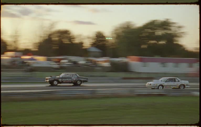 A pair of cars drag race down a race track in the evening