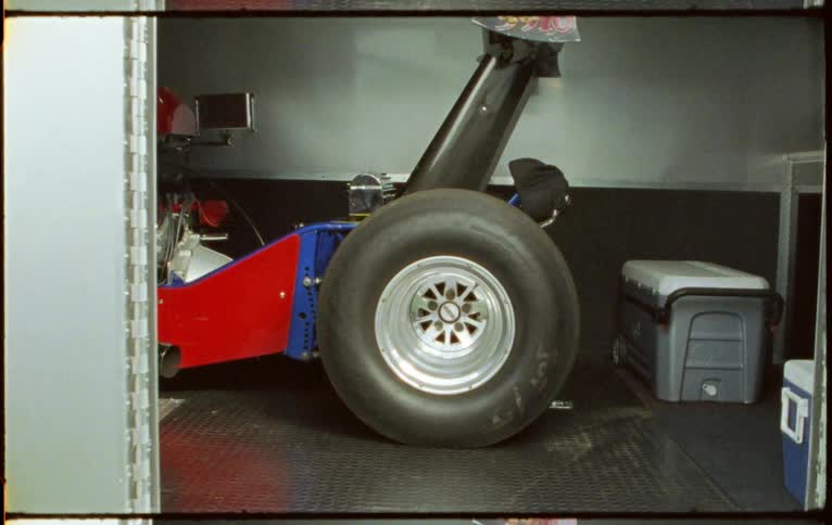 A drag racing car with a huge rear tire is parked inside of a transportation trailer