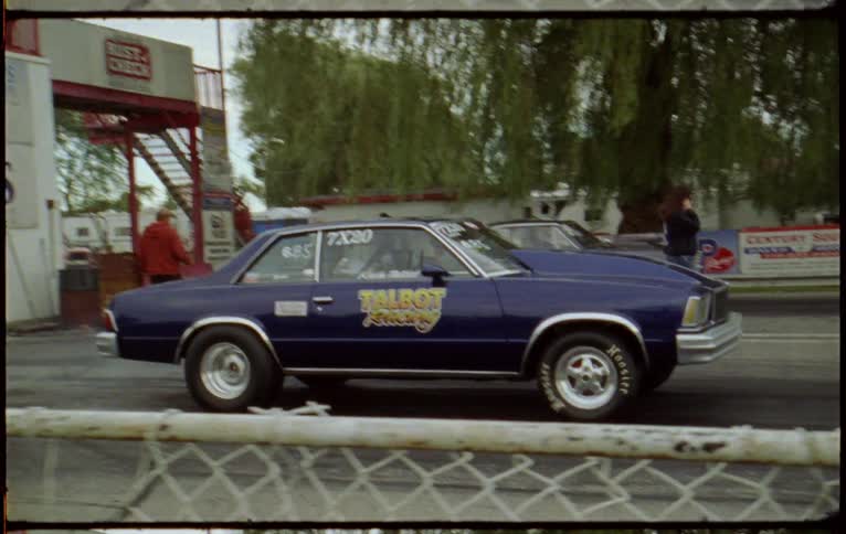 Two vehicles drag racing on a track with trees the sky and clouds in the background
