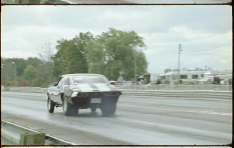 Sports cars race down track past parking lot and woods