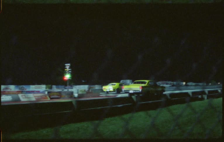 Two vehicles on a racetrack drag racing with grass and a fence in the foreground