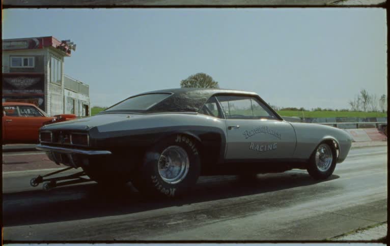 A red sports car and a gray muscle car speed off as they race on a straight track