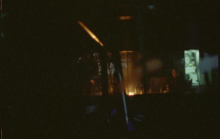 A small fire burns outside of a building as silhouettes of people can be seen