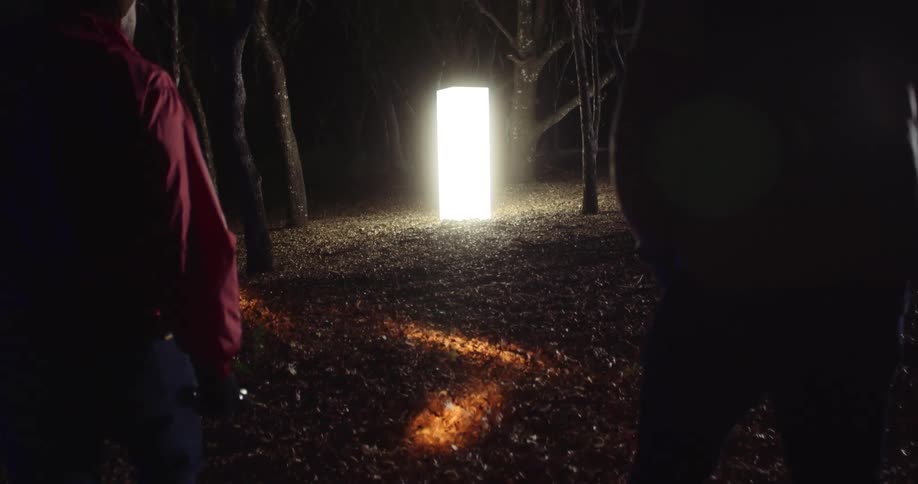 Three people walk through a wooded area at night with flashlights towards a bright lamp structure