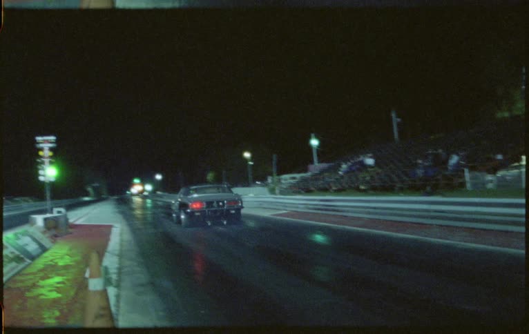 A vehicle driving on a racetrack with bleachers and streetlights in the background