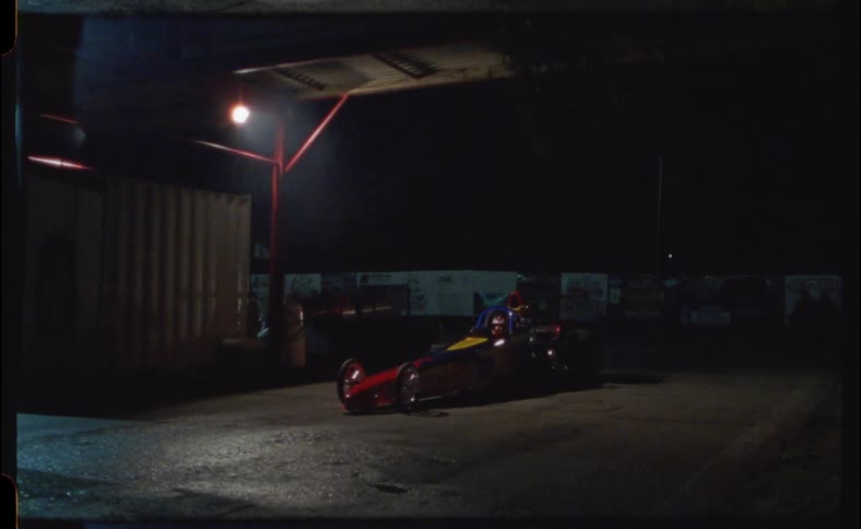 A race car with an elongated pointed front pulls up to a building