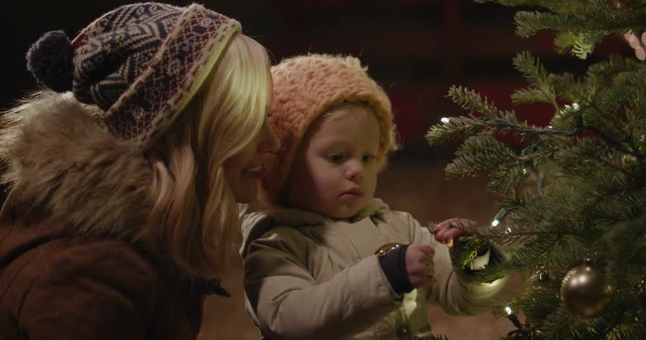 A mother talks to her toddler who is placing an ornament on a christmas tree
