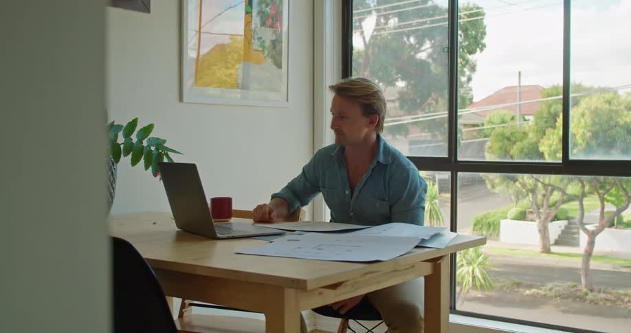 A man in a room sitting on a chair at a table using a laptop
