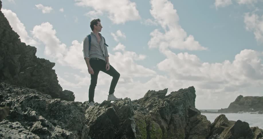 A man wearing a backpack while standing on a rock formation on the coast overlooking the ocean gazes around at his surroundings