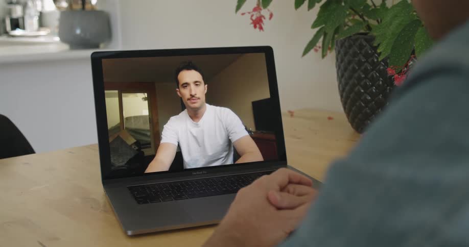 A man on screen on a laptop talks to another sitting at a table with hands folded