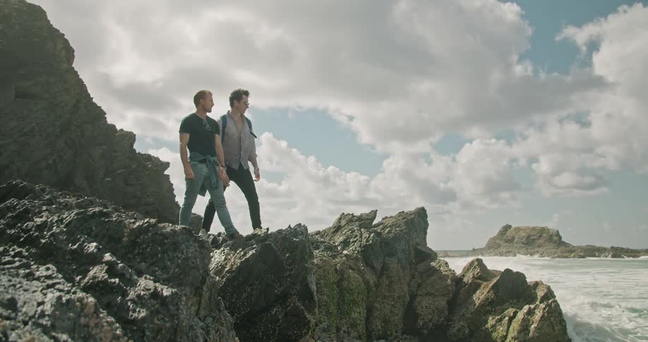 A pair of men stand together on a rock formation overlooking the ocean on the coast during the day