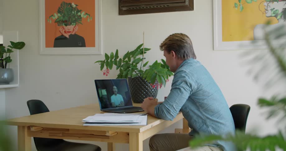 A man sits at a desk in his home office talking to another on a video call meeting
