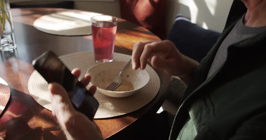 A man sitting a dining table in a house shows a text message on his smartphone to a woman sitting beside him