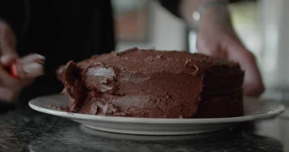 Man spreads chocolate icing on chocolate cake standing in kitchen