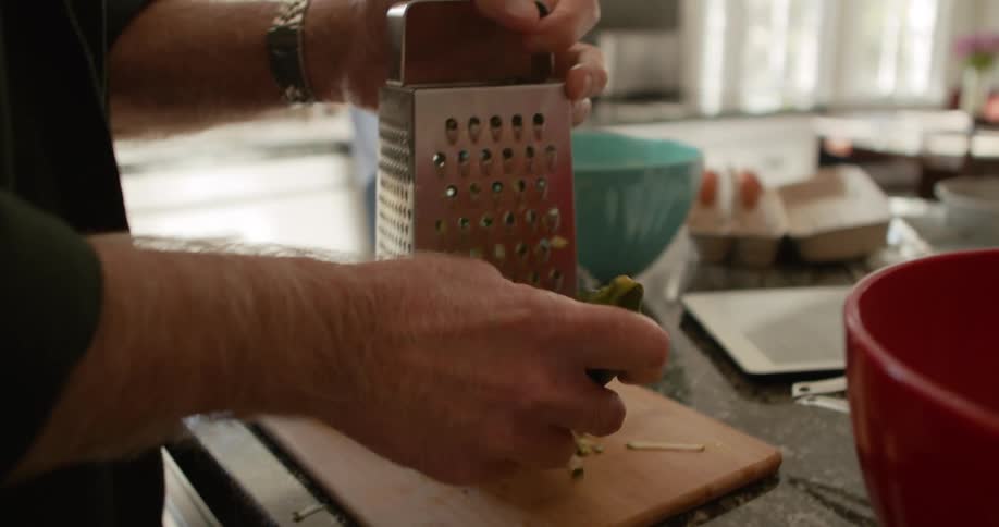 An older man carefully uses a grater to grate a single zucchini in his kitchen